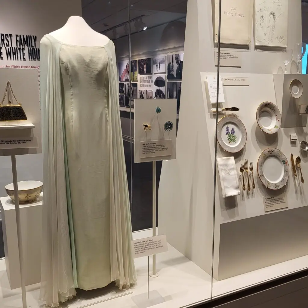A display showcasing a formal gown worn by First Lady Lady Bird Johnson, along with official White House china and place settings in an exhibit at the LBJ Presidential Library in Austin, Texas